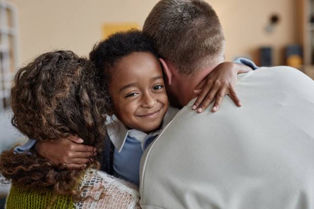 Portrait shot of little happy Black boy giving tender embrace to mother and father while supporting parents caring for adopted kid in childs bedroom